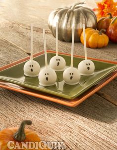 four white cake pops with ghost faces on them sitting on a green plate next to pumpkins