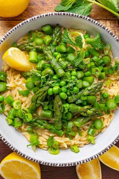 a white bowl filled with pasta and peas next to lemon wedges on a wooden table