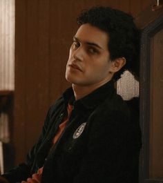 a young man with curly hair sitting in front of a wooden wall and looking off into the distance