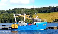 a blue fishing boat in the water next to a dock with trees and hills in the background