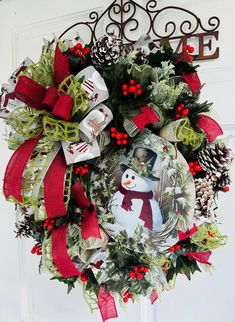 a christmas wreath with a snowman on it hanging from the front door, decorated with red ribbon and pine cones