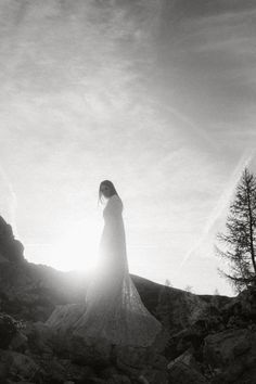 a woman standing on top of a rocky hillside under a cloudy sky with the sun shining through