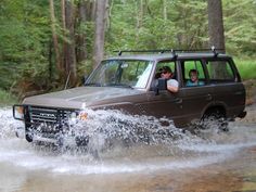 two people are driving in the water behind a land rover on a muddy road near some trees