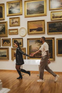 a man and woman holding hands while walking in front of paintings on the wall behind them