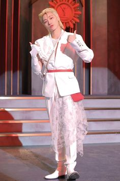 a woman in a white suit and red belt is giving the thumbs up while standing on stage