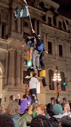 two men standing on top of a traffic light while holding onto the same flag as others watch