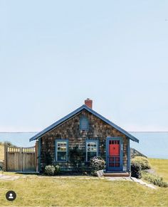 a small house sitting on top of a lush green field next to the ocean with a red door