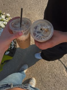 two people holding drinks in their hands on the ground with white flowers behind them and one person's legs showing