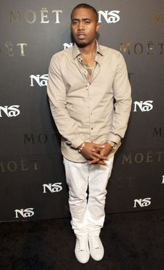 a man standing in front of a black wall wearing white pants and a tan shirt