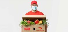 a man wearing a face mask while holding a cardboard box filled with vegetables and fruits