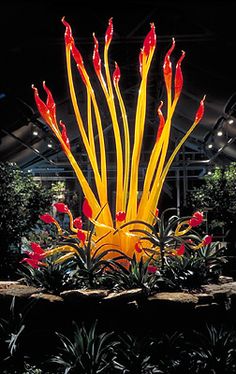 a large yellow sculpture with red flowers in the middle of some trees and plants around it