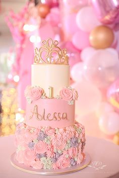 a pink and gold birthday cake with flowers on the bottom, sitting on a table in front of balloons