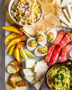 an assortment of food is laid out on a marble table with chips, guacamole, and fruit