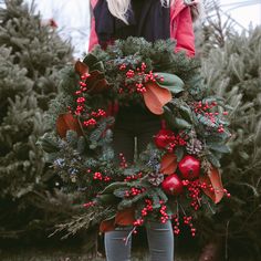 a woman holding a christmas wreath with berries and pine cones on the front, surrounded by evergreens