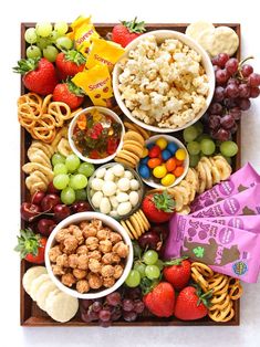 a wooden tray filled with fruit and snacks next to crackers, pretzels, grapes, strawberries