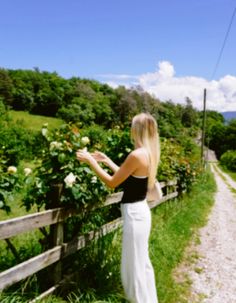 a woman standing next to a wooden fence in front of green grass and flowers on the other side