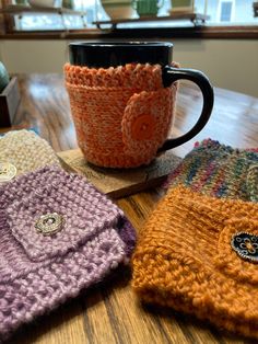 three knitted coffee mug cozyies sitting on top of a wooden table next to a cup