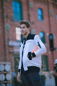 a young man standing in front of a brick building wearing a white and black jacket