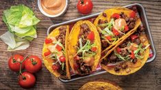 three tacos on a tray with tomatoes, lettuce and sauce next to them