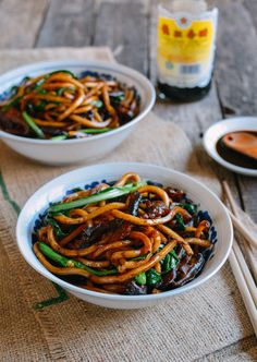 two white bowls filled with stir fry noodles on top of a wooden table next to chopsticks