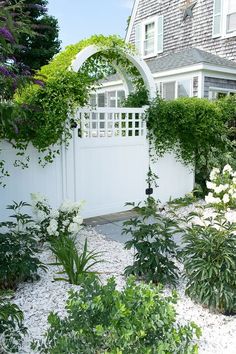 a white gate surrounded by plants and flowers in front of a house with an arched window