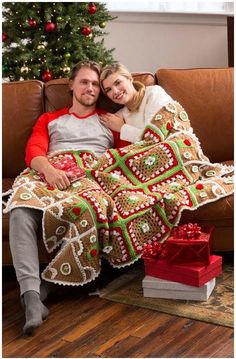 a man and woman sitting on a couch under a christmas tree with a blanket draped over them