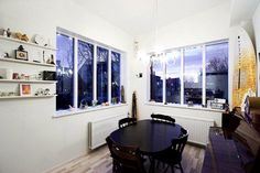 a dining room table and chairs in front of two large windows with open shelves on the wall