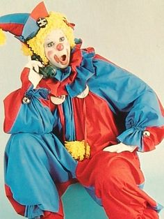 a clown is sitting on the ground with his hands in his pockets and mouth open