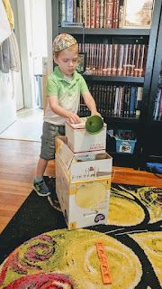 a young boy standing on top of a cardboard box filled with green apples and cucumbers