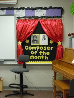 a classroom with a piano, desk and red curtains on the wall above it is a poster that says composer of the month