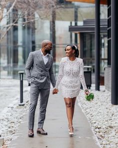 a man and woman are walking down the sidewalk holding hands while dressed in grey suits