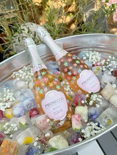 two bottles of champagne sitting on top of ice cubes in a metal bowl filled with flowers