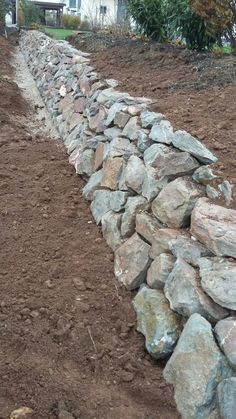 a stone wall in the middle of a field with dirt and rocks on it, next to a house