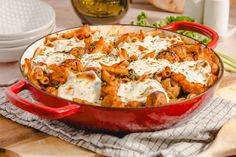 a red casserole dish filled with pasta and sauce on a wooden cutting board