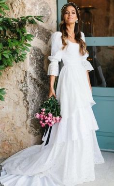 a woman in a white wedding dress standing next to a stone wall holding a bouquet of flowers