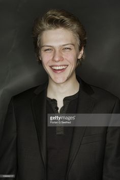 a smiling young man in a suit and black shirt, with his hair pulled back