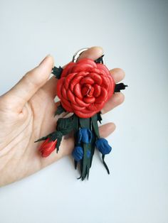 a hand holding a red rose brooch with green leaves and blue flowers on it