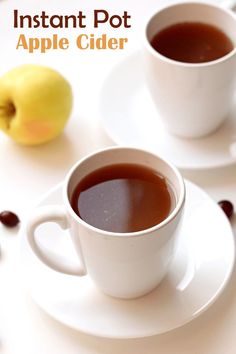 two white cups filled with coffee sitting on top of a plate next to an apple