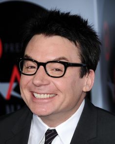 a man wearing glasses and a suit smiles for the camera while standing in front of a red carpet