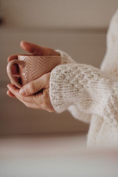 a woman holding a coffee cup in her right hand while wearing a white knitted sweater