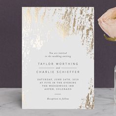 a white and gold wedding card next to a vase with a rose in it on a marble surface