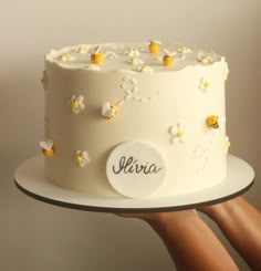 a hand holding a white cake with daisies on it and the name shrira