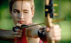 a young woman is aiming an arrow with her right hand and looking at the camera