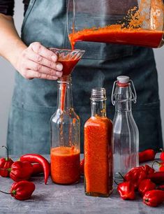 a person pouring red sauce into a glass bottle next to bottles with chili peppers on the table