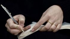 a person holding a pen and writing on an open book with two gold rings around their fingers