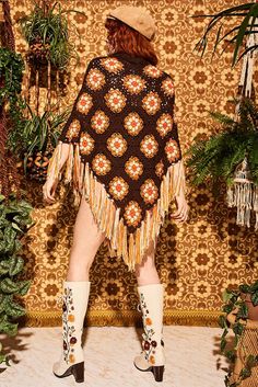 a woman standing in front of a wall covered in plants and wearing a brown poncho