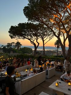 many people are sitting at tables with food on them in front of some trees and the sun is setting