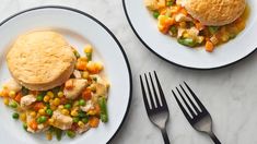 two plates filled with food next to silverware on a white counter top, one has a biscuit and the other has vegetables