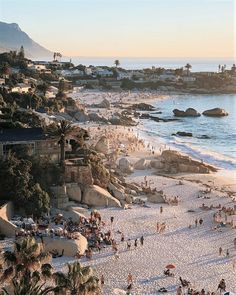 many people are on the beach and in the water near some rocks, palm trees and mountains