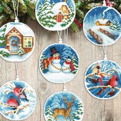 cross stitch christmas ornaments hanging on a wooden table with pine branches and evergreens in the background